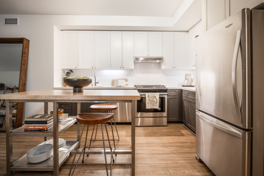 Open style kitchen with grey cabinets on the bottom and white cabinets on the top. White countertop, and stainless steel appliances.